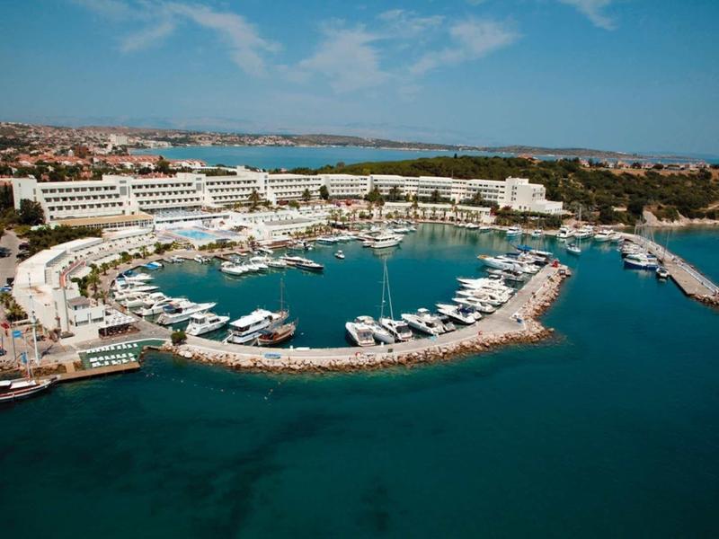 Hafen mit Yachten, weißem Hotelkomplex und blauem Meer unter klarem Himmel.