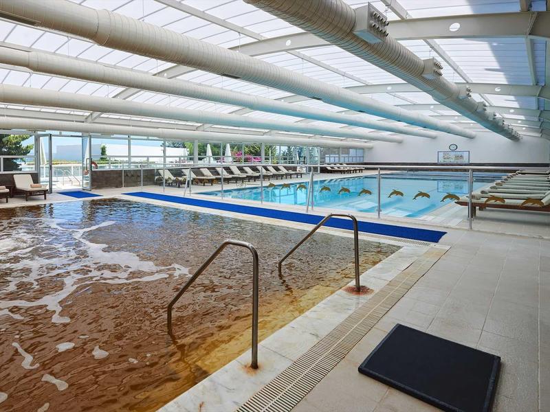 Innen-Schwimmbad mit zwei Becken, Liegestühlen und bodentiefen Fenstern für natürlichen Lichteinfall.