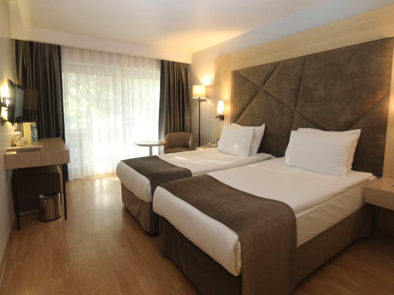 Modernes Hotelzimmer mit zwei Betten, hellen Holzböden und großem Fenster mit Vorhängen.