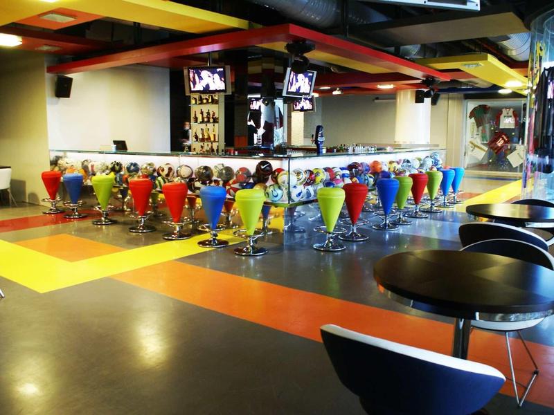 Großer Raum mit bunten Stühlen, schwarzen Tischen und zwei Fernsehern an der Decke.