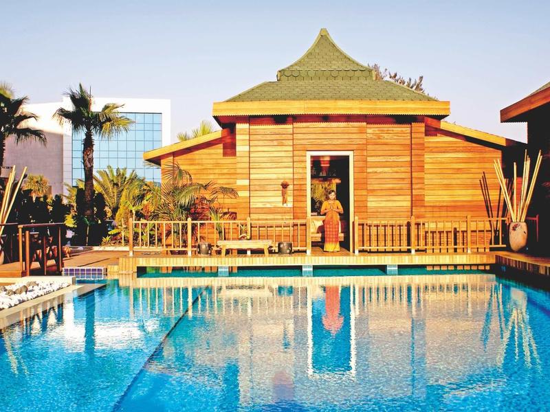 Blauer Pool mit hölzerner Hütte, umgeben von Palmen und Gebäuden bei klarem Himmel.