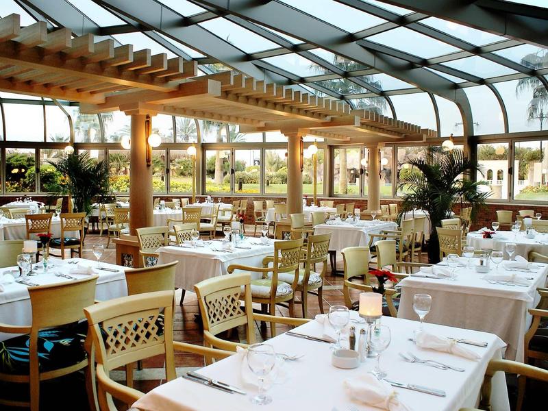 Großes Restaurant mit Glasdach, Holzbalken, weißen Tischdecken und Holzstühlen mit gepolstertem Sitz.