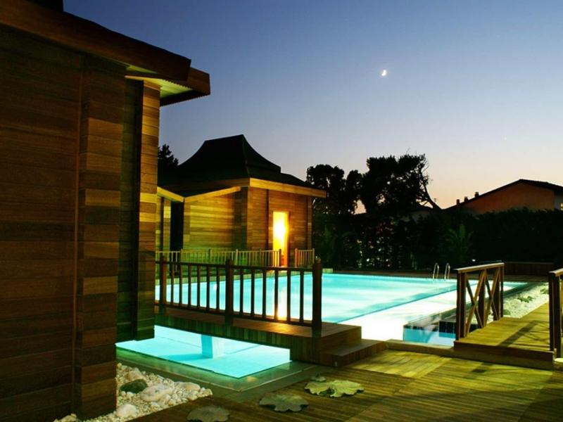 Moderner beleuchteter Pool mit Holzhaus und Terrasse bei Dämmerung und klarem Himmel.