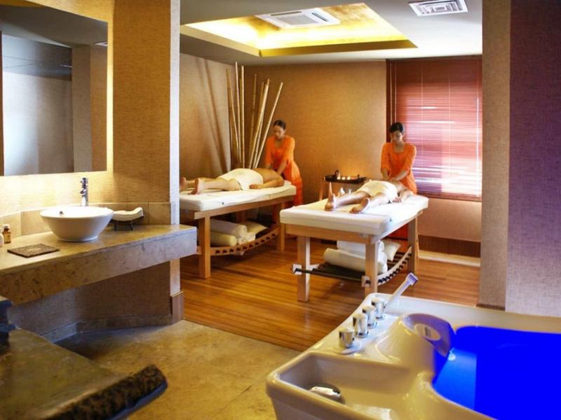 Wellnessraum mit Massageliegen, Badewanne, warmem Licht und zwei Personen in orangefarbenen Bademänteln.