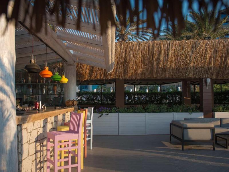 Outdoor-Barbereich mit pinken Barhockern, Strohdach und moderner Lounge-Möblierung bei Sonnenuntergang.