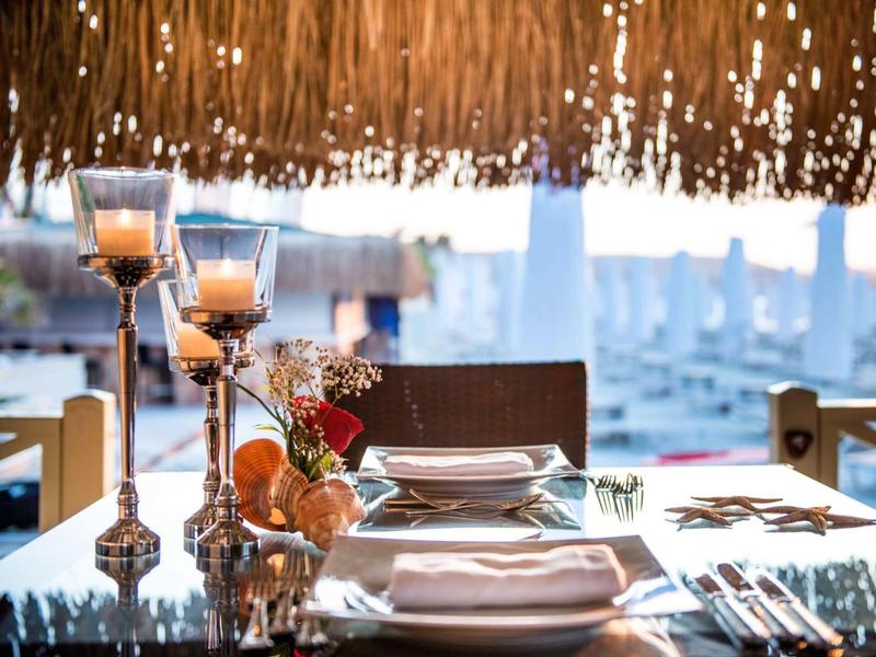 Gemütlicher gedeckter Tisch mit Kerzen unter Strohdach, im Hintergrund Boote im Hafen sichtbar.