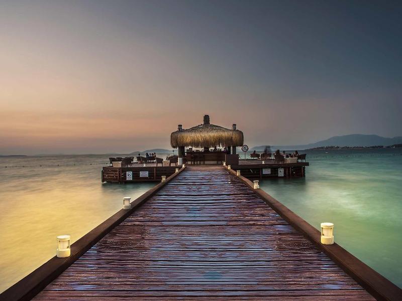 Ein langer hölzerner Steg führt zu einer überdachten Plattform über ruhigem Wasser bei Sonnenuntergang.