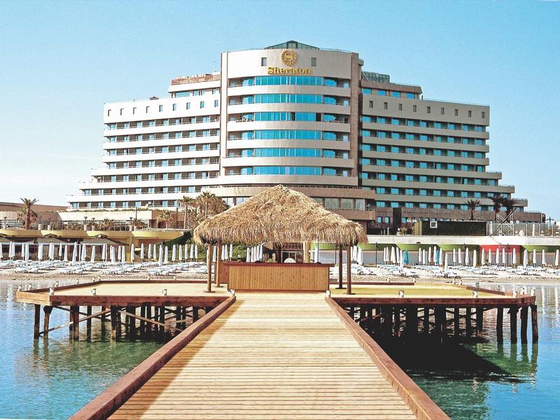 Großes weißes Hotel am Wasser mit Holzsteg und Strohdach-Pavillon im Vordergrund.