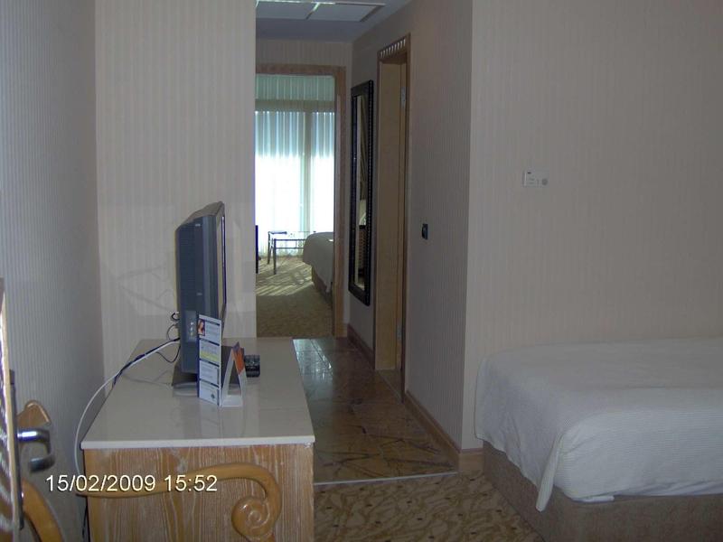 Hotelzimmer mit Bett rechts, Fernseher auf Schreibtisch links und Flur zu weiterem Raum im Hintergrund.