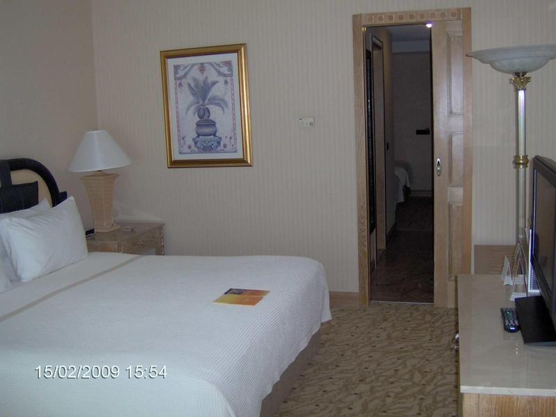 Ein schlichtes Hotelzimmer mit großem weiß bezogenem Bett, Nachttisch, Lampe und Bild an der Wand.