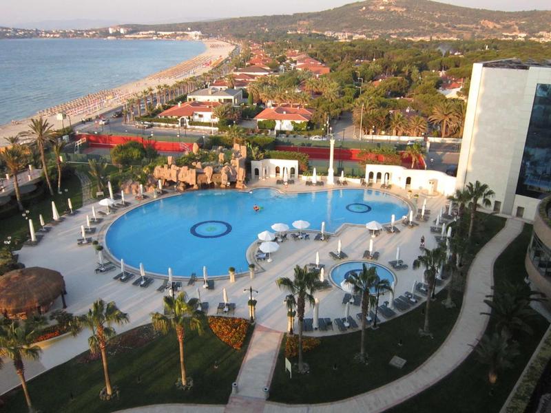 Großer runder Pool mit weißen Liegen und Sonnenschirmen, umgeben von Palmen an der Küste.