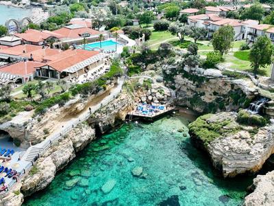 Küste mit klarem, türkisfarbenem Wasser, Felsen, Sonnenliegen, Bäumen und Gebäuden im Hintergrund.