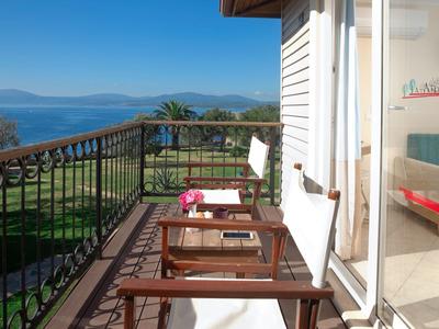 Balkon mit Holzstühlen, Metallgeländer, Blick auf Meer und Berge bei klarem Himmel.