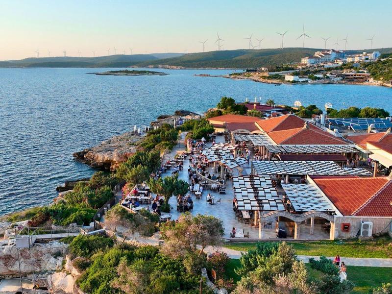 Küstenrestaurant mit roten Dächern, Terrasse voller Menschen, Blick auf ruhiges Meer und grüne Hügel im Hintergrund.