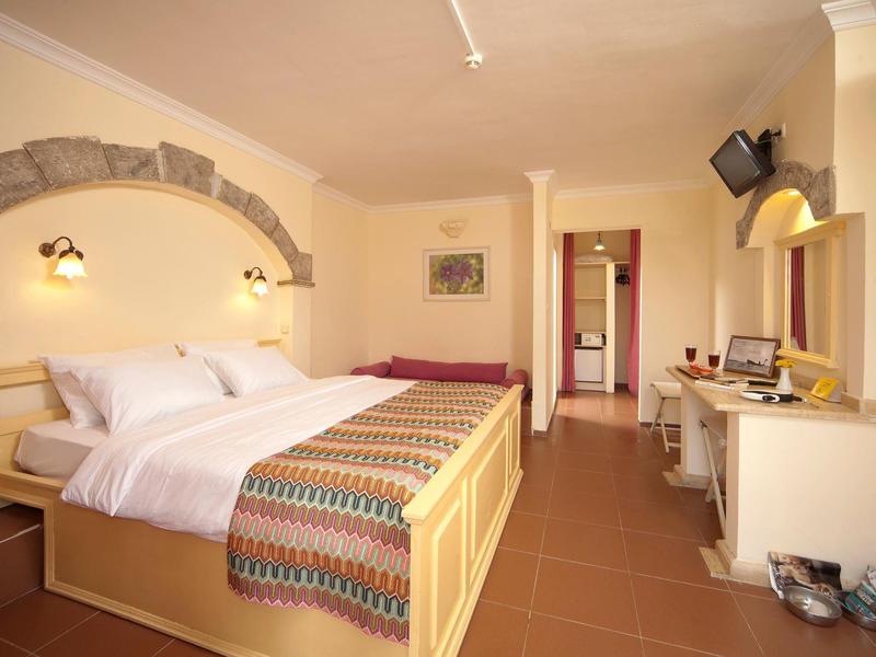 Helles Schlafzimmer mit Doppelbett, buntem Bettläufer, Schreibtisch, Stuhl und kleinem Fernseher an der Wand.