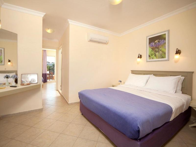 Helles Hotelzimmer mit großem Bett, blauer Decke, Fliesenboden und Wandbild über Kopfteil.