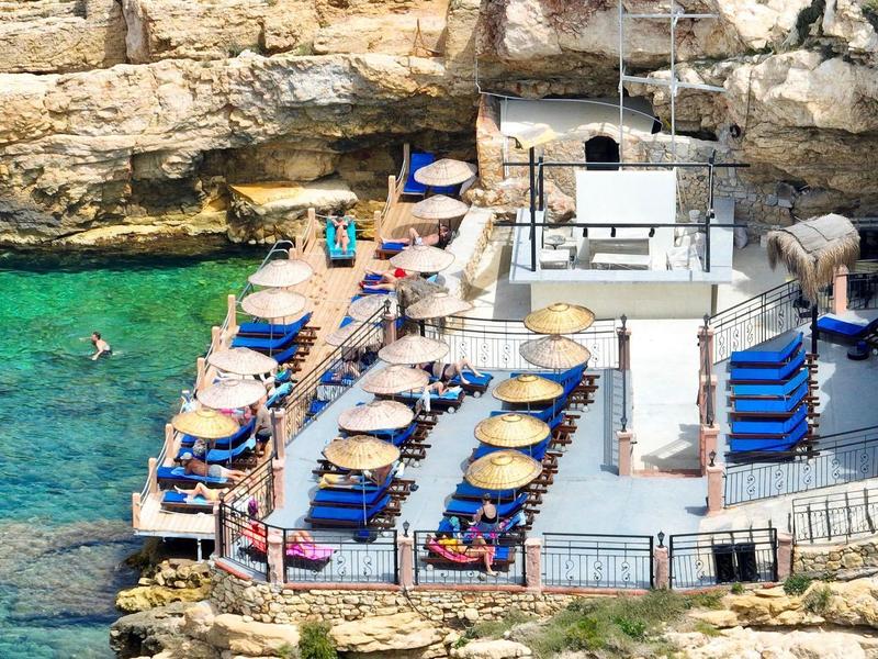 Eine Terrasse mit Sonnenschirmen und Liegestühlen am türkisfarbenen Meer, umgeben von Felsen.