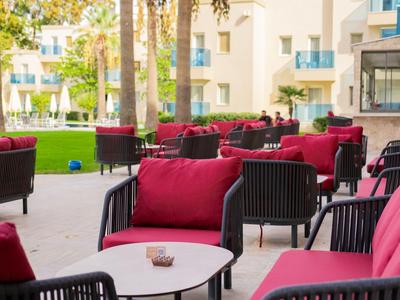 Outdoor-Loungebereich mit roten Sitzkissen und Tischen vor einem Hotel mit Palmen und Garten.