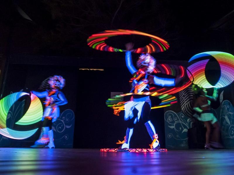 Drei Performer mit leuchtenden, bunten LED-Reifen tanzen auf dunkler Bühne.