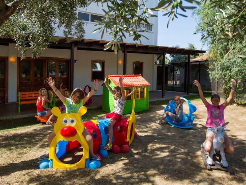 Kinder spielen draußen auf buntem Spielzeug und Schaukeltieren im sonnigen Garten vor einem Haus.