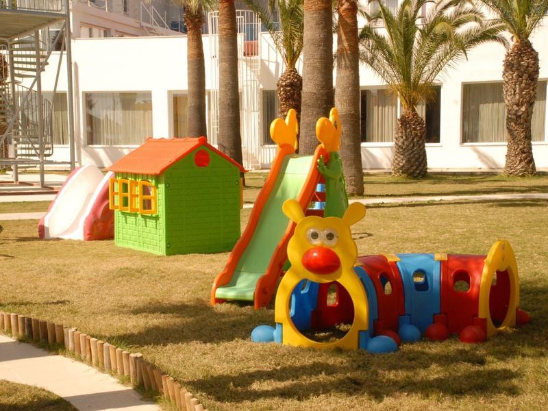 Bunter Spielplatz mit Rutsche, Spielhaus und Raupen-Tunnel auf Grasfläche neben Palmen.