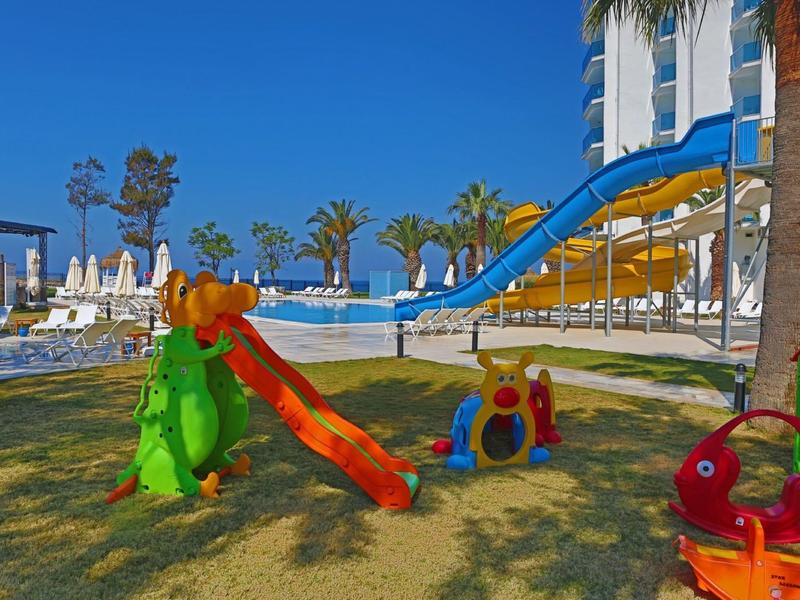 Bunter Kinderspielplatz mit Rutschen und Schaukel im Sonnenschein, Palmen und Hotel im Hintergrund.
