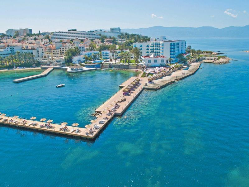 Küstenstadt mit Hafen, blauer See, Liegeplätzen, weißen Gebäuden und grünen Hügeln im Hintergrund.