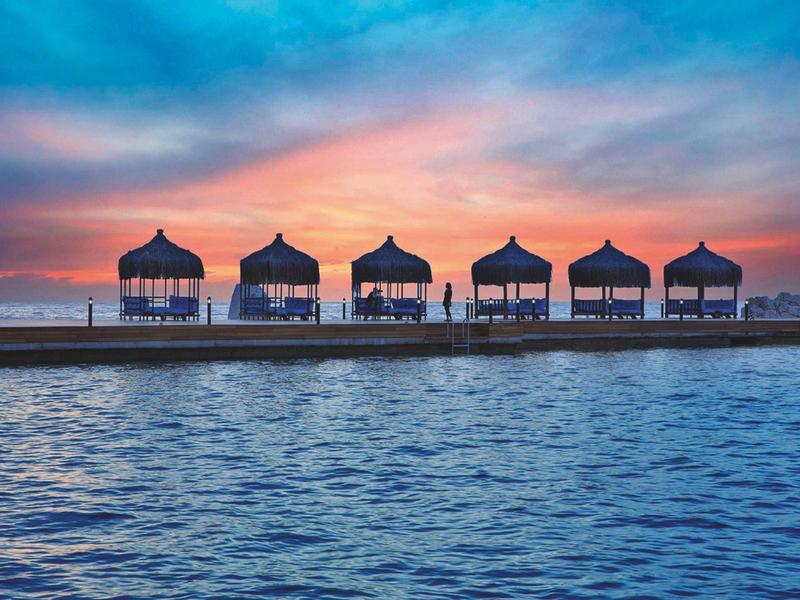 Sechshäusige Strandpavillons bei Sonnenuntergang über ruhigem Meer mit farbigem Himmel.
