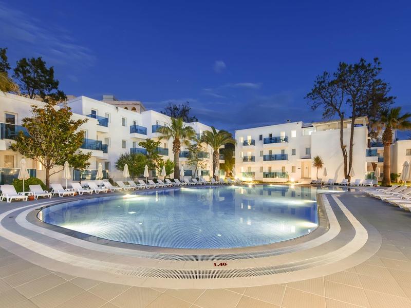 Großer beleuchteter Pool vor einem modernen Hotelgebäude mit Liegestühlen und Palmen bei Abenddämmerung.