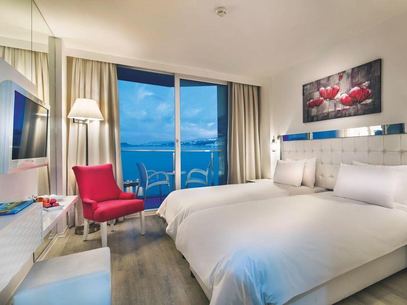 Helles Hotelzimmer mit großem Bett, rotem Sessel, Balkon und Meerblick.