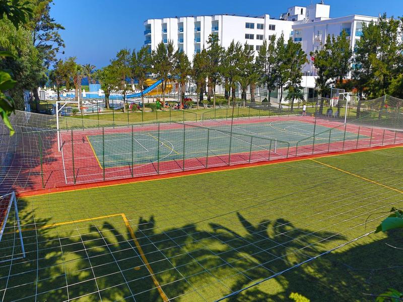 Ein eingezäunter Tennisplatz mit rotem Boden neben einem grünen Rasen und weißen Gebäuden im Hintergrund.