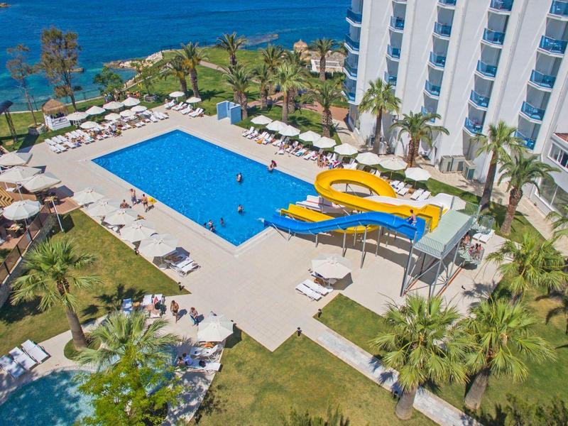 Hotel mit großem blauen Pool, gelber Wasserrutsche, Palmen und Liegestühlen am Meerufer.