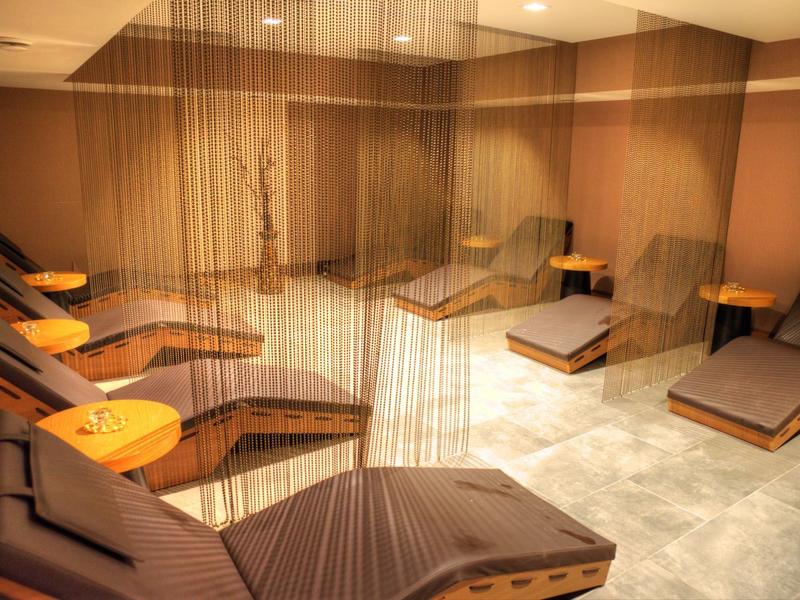 Ruheraum mit braunen Liegen, warmem Licht und zarten Vorhängen aus Fäden für entspannte Atmosphäre.