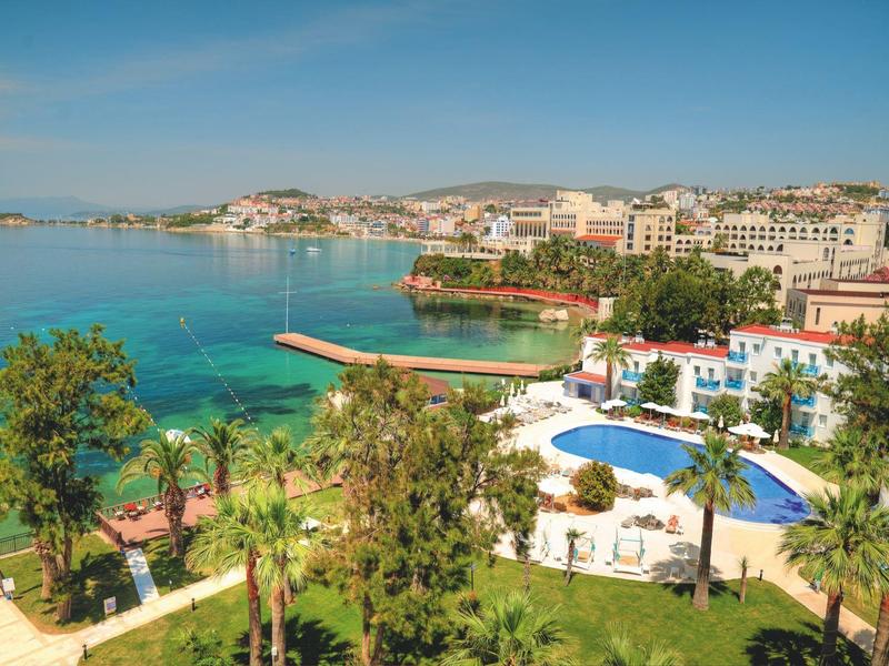 Klares blaues Meer, Pool mit Sonnenschirmen, grüne Palmen und helle Gebäude am Küstenhügel.