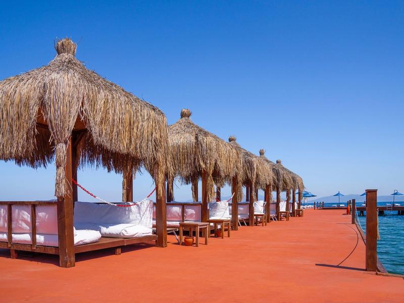 Rote Holzterrasse mit Strohdächern über Liegen, daneben blaues Meer und klarer Himmel.