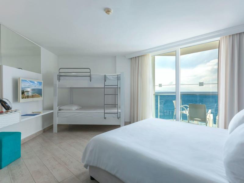 Helles Schlafzimmer mit Doppelbett, Etagenbett, großem Fenster und Zugang zu Balkon mit Meerblick.