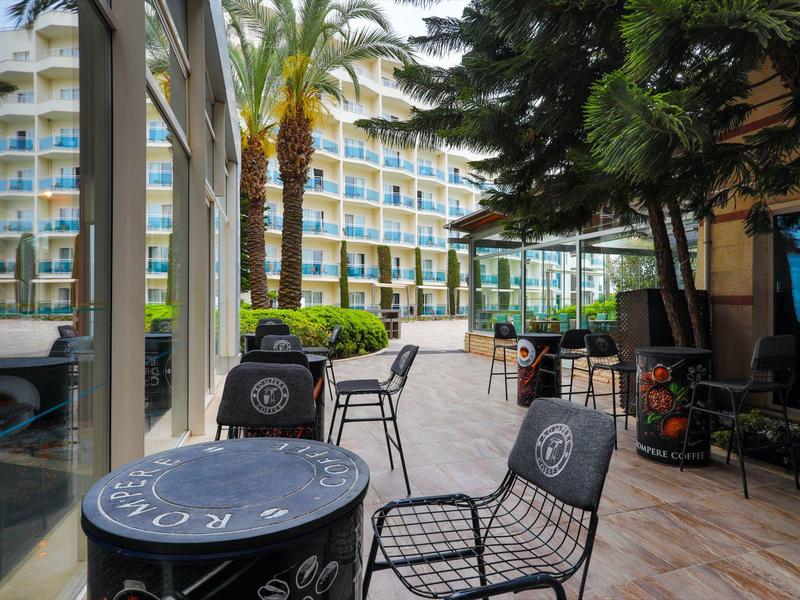 Outdoor patio area with metal tables and chairs surrounded by palm trees near a hotel building.