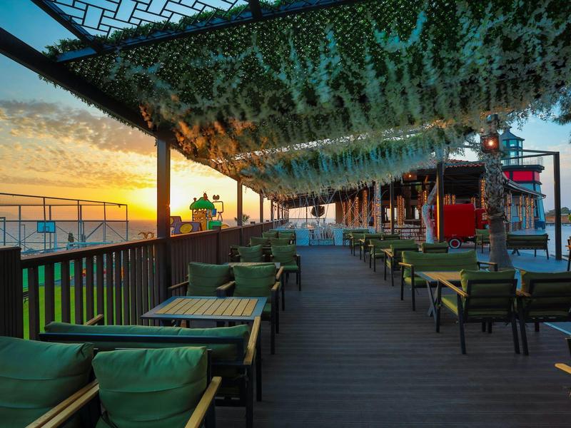Outdoor hotel terrace with green cushions, tables, and overhead leafy canopy at sunset by the sea.