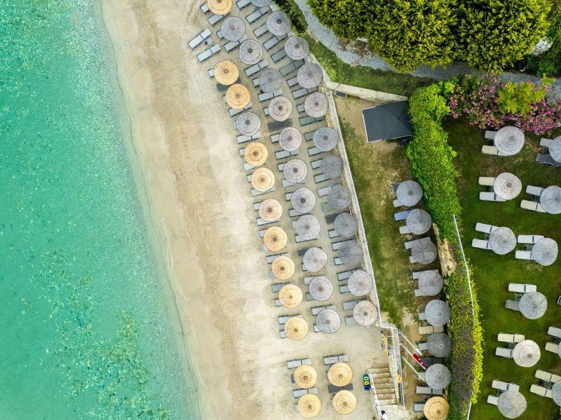 Aerial view of a beach with turquoise water, umbrellas, sunbeds, and green garden areas.