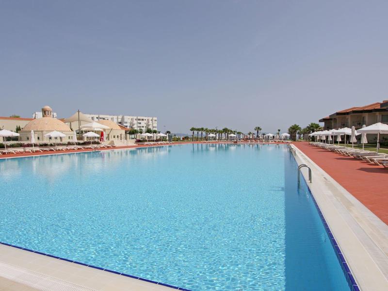 Großer, klarer Swimmingpool mit blauer Wasseroberfläche, umgeben von Liegestühlen und Gebäuden.