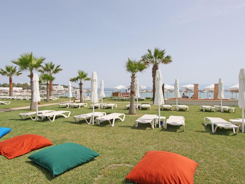 Grüner Rasen mit bunten Kissen, weißen Liegen, Palmen und blauem Himmel am Meer.