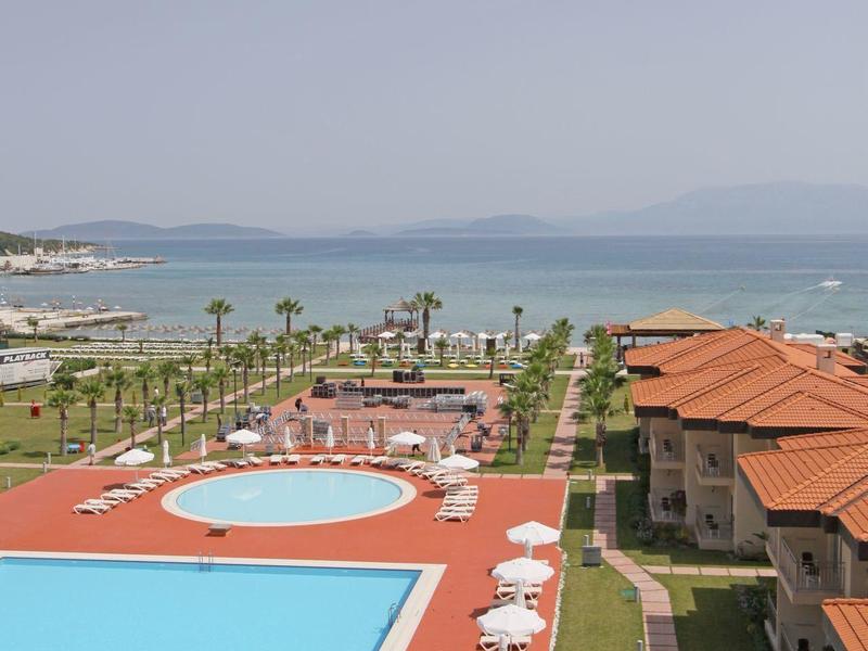 Großer Pool mit weißen Sonnenschirmen, roten Wegen, grünen Palmen und Meer im Hintergrund.