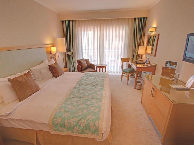 Modernes Hotelzimmer mit großem Bett, Schreibtisch, TV und Balkon hinter weißen Vorhängen.