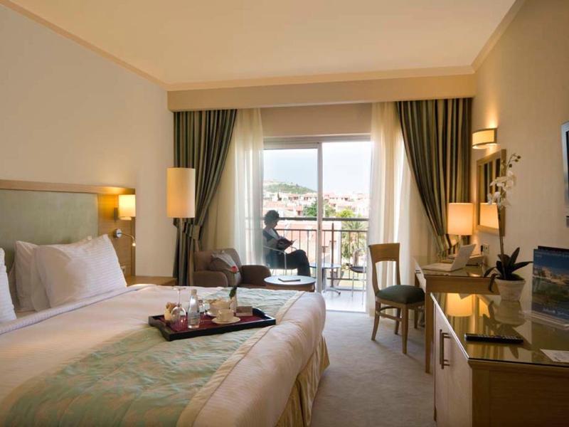 Helles Hotelzimmer mit Doppelbett, Balkon, zwei Sesseln und Schreibtisch mit Fernseher.