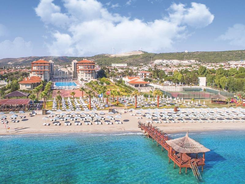 Strand mit klarem blauem Wasser, Liegen, Sonnenschirmen und einem Steg zum Meer vor einem Hotelkomplex.