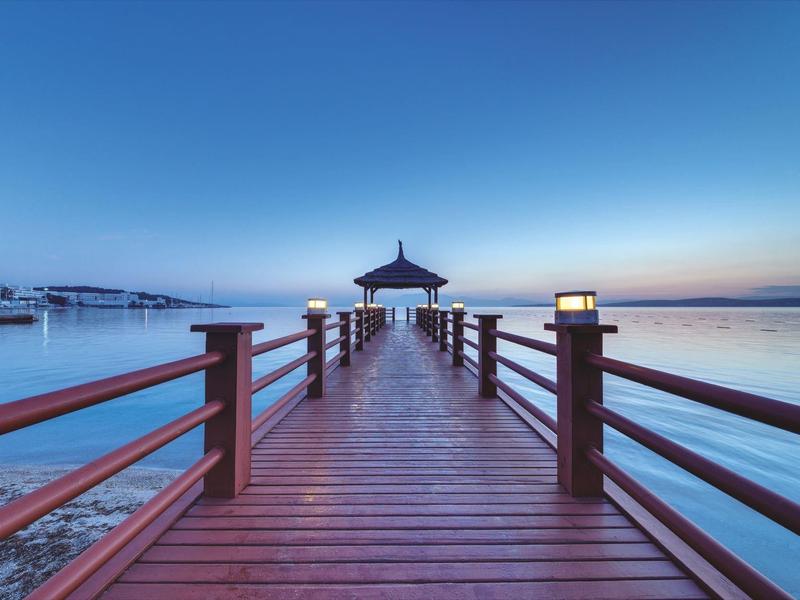 Langer Holzsteg mit Laternen führt über ruhiges Wasser zum Pavillon bei blauem Himmel.