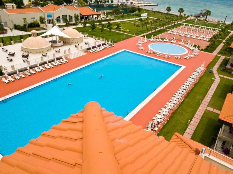 Großer blauer Pool mit weißen Liegestühlen umgeben von grünen Rasenflächen und orangefarbenen Dächern.