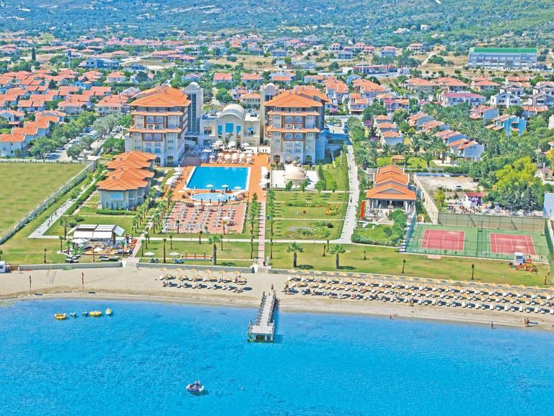 Große Hotelanlage mit mehreren Gebäuden, Pool, Tennisplätzen und Strand am blauen Meer.