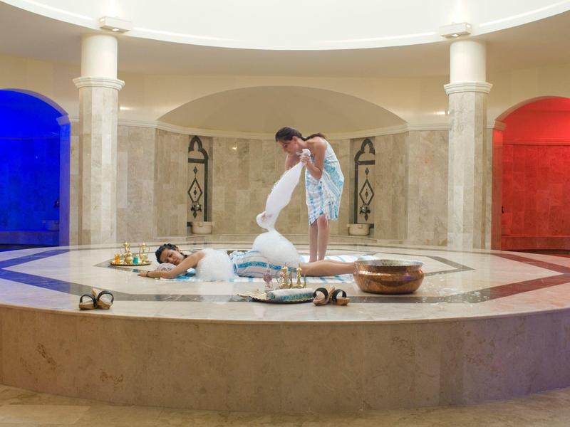 Ein Mann massiert eine liegende Person in einem runden Raum mit Mosaikboden und blau-roten Wänden.