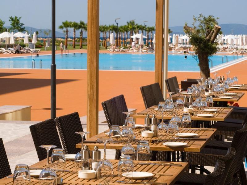 Outdoor-Restaurant mit Holztischen und Stühlen, Blick auf blauen Pool und Himmel im Hintergrund.